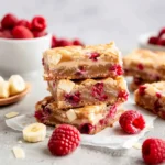 Close-up of raspberry and white chocolate blondies showing fudgy center and crackly golden top with jammy raspberry pockets and melted white chocolate on a wooden board.