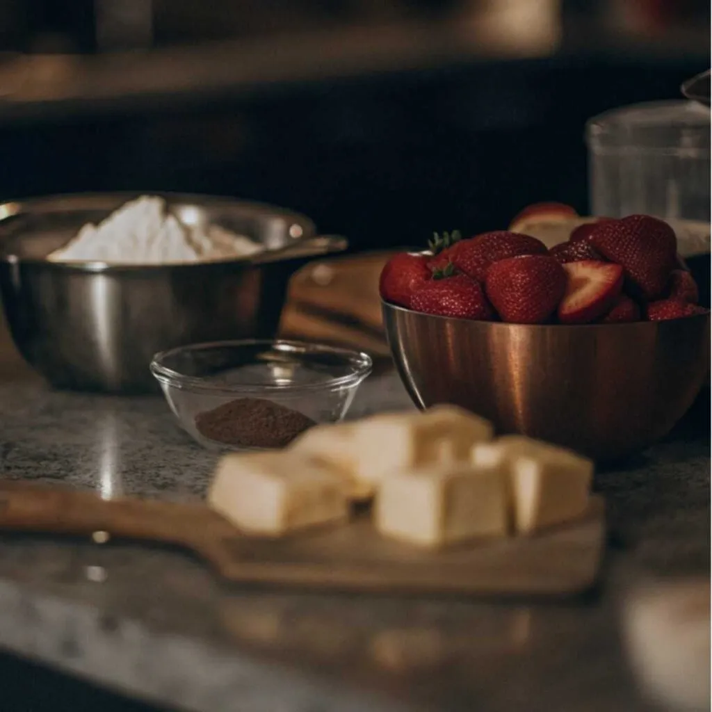 Ingredients for strawberry shortcake including fresh strawberries, flour, and butter arranged on a dark kitchen countertop.