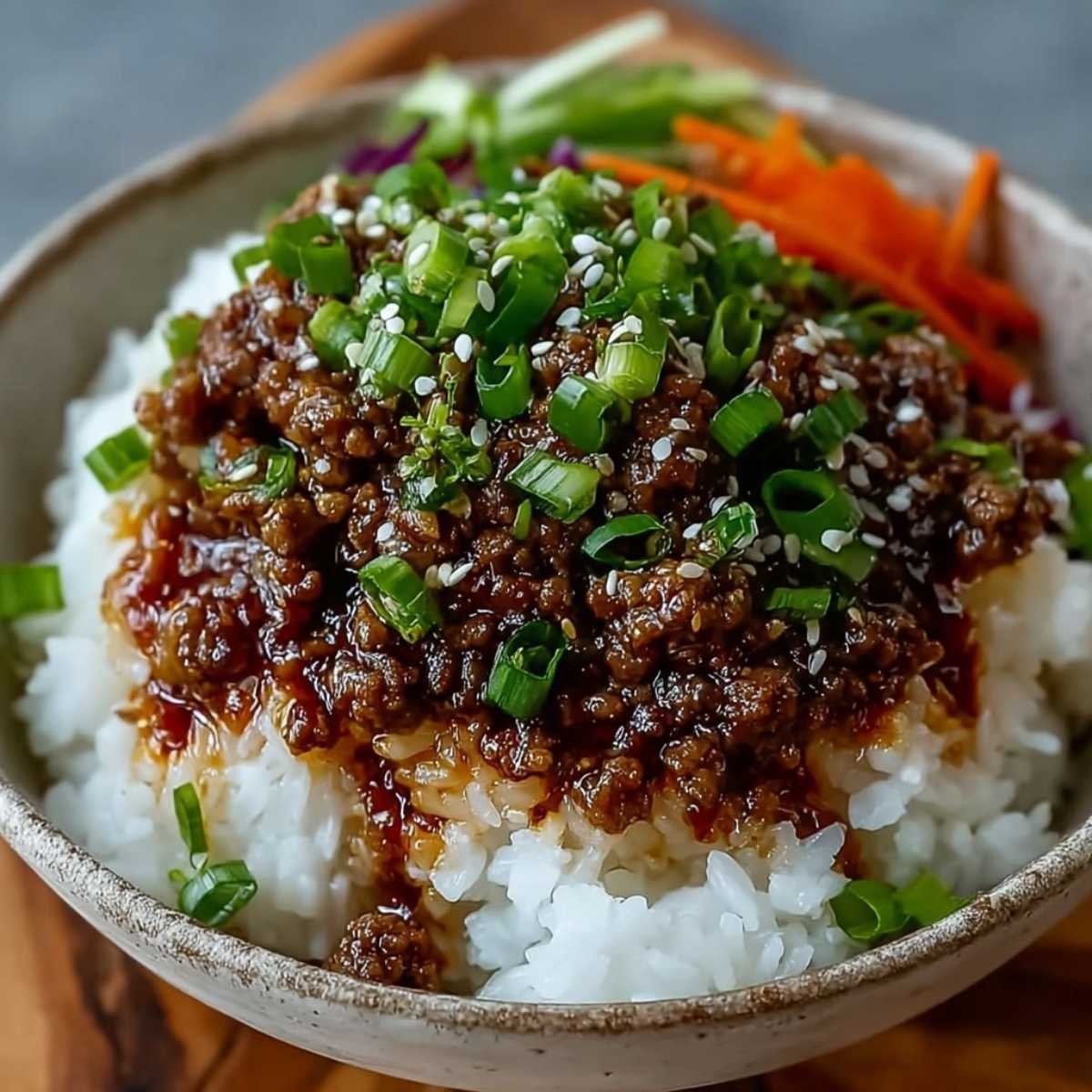 Close-up of Easy Korean Ground Beef Bowl with sweet-savory beef over steamed white rice, topped with fresh green onions and sesame seeds, with shredded carrots, cucumber, and cabbage in the background