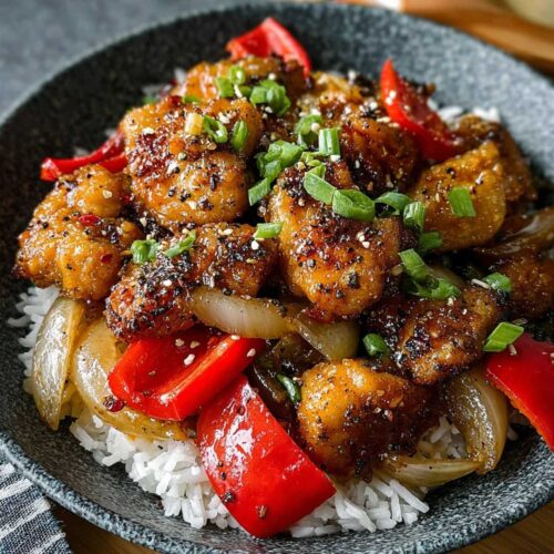 Black Pepper Chicken in a dark grey bowl, showcasing its rich, peppery sauce, red bell peppers, and fresh green onion garnish over white rice