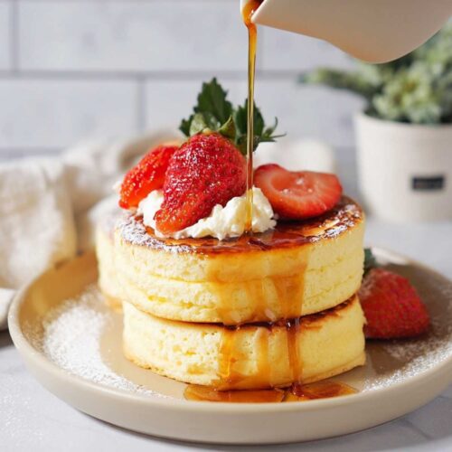 Japanese Soufflé Pancakes on a modern grey plate, showcasing their towering height with fresh strawberries and maple syrup drizzled on top.