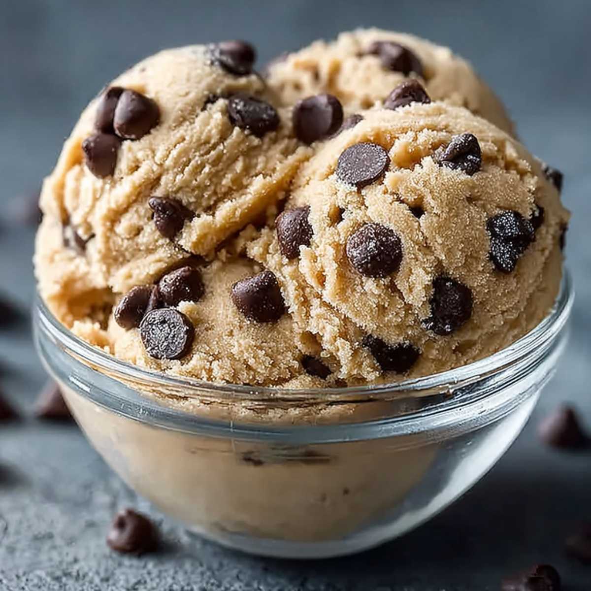 Three scoops of protein cookie dough in a clear glass bowl, showcasing chocolate chips and creamy texture on a gray surface